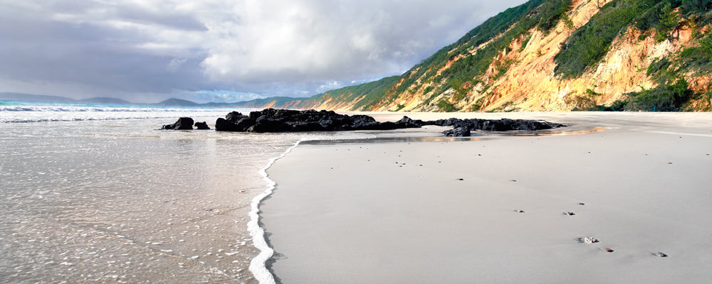Dark rocks on wet sand below the coloured cliffs of Rainbow Beach, Queensland - fine art photography print by Adam Gormley